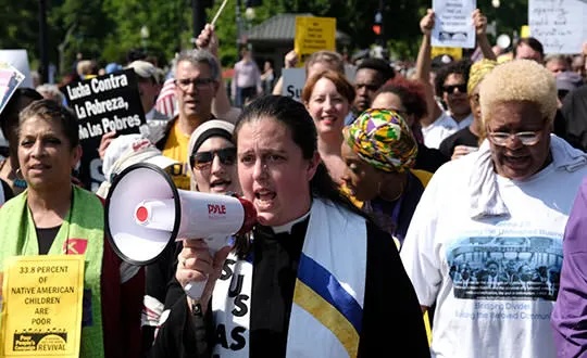Liz Theoharis speaks in bullhorn at a rally amidst a crowd of people holding signs calling for human rights for the poor